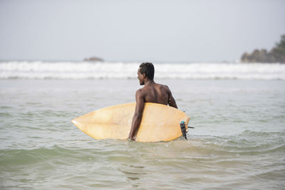 Rear view of shirtless male surfer carrying surfboard in sea