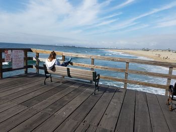 Rear view of woman sitting on bench at pier over sea against sky