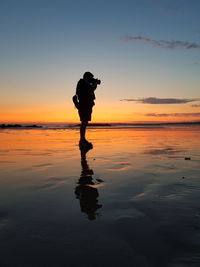 Silhouette man photographing while standing at beach against sky during sunset