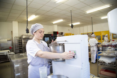 Focused female worker in uniform turning on modern dough mixer with metal bowl while working in professional light bakery with colleagues