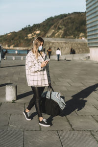 Woman in face mask using smart phone while walking on footpath in city