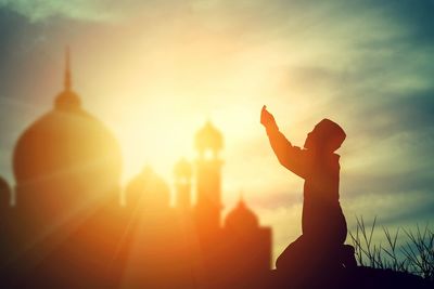 Silhouette boy praying against mosque during sunset