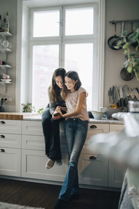 Full length of smiling female friends using mobile phone in kitchen at home