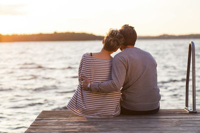 Rear view of affectionate couple sitting on pier against sea at sunset