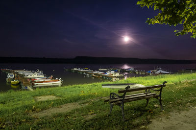 Empty bench on grass by lake against sky at night