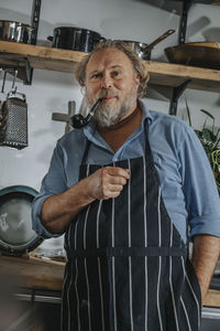 Mature chef wearing apron standing with pipe and hands in pockets at kitchen