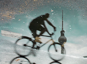 Reflection of man riding bicycle with fernsehturm in puddle on street