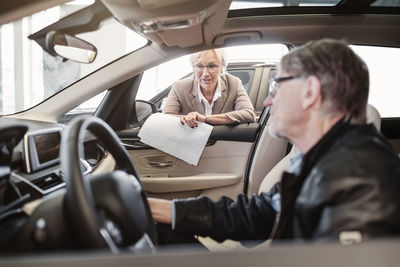 Senior saleswoman explaining features to man sitting in car at shop