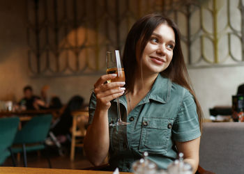 Beautiful embarassed female with cheerful expression sitting with glass of wine in the cafe 