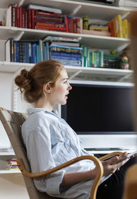 Side view of woman holding digital tablet while sitting on chair at home