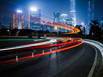 Light trails on city street by buildings against sky at dusk