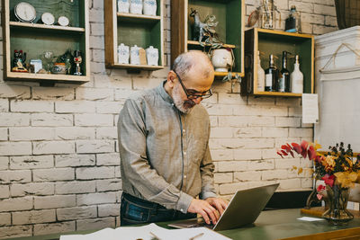 Senior male owner working on laptop at checkout counter in interior shop