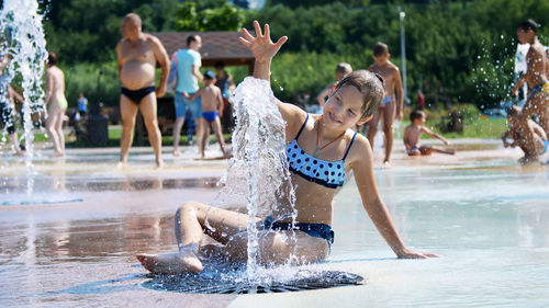 Smiling, happy eight year old girl in swimsuit having fun in splashes in street city fountain
