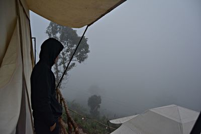 Side view of man standing by tent during foggy weather against sky