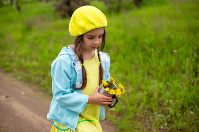 A little girl walks along a path near a green lawn with a bouquet of yellow dandelions in an mug