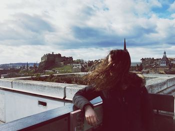 Rear view of woman sitting on retaining wall in city against sky