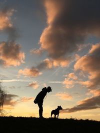 Silhouette woman standing with dog on field against sky during sunset