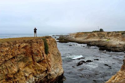 Man standing on cliff by sea against sky