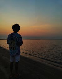 Rear view of man walking at beach against sky during sunset