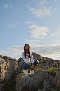 Full length of young woman crouching on rock against sky