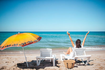 Rear view of woman sitting on beach against clear blue sky