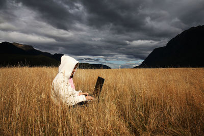 Mid adult woman using laptop while sitting on grassy field against cloudy sky