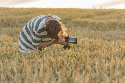 A teenager in a striped t-shirt shoots a sunset on the phone in a wheat field.