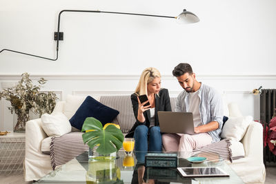 Positive blond woman with cup of juice browsing smartphone and sitting on couch near ethnic boyfriend typing on laptop keyboard in living room of modern apartment