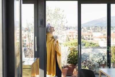 Senior woman with coffee cup wrapped in blanket leaning on sliding door at home