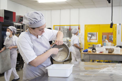 Side view of female baker in uniform putting raw dough from bowl into container while working in professional light bakery