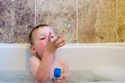 Playful boy holding soap bubble while sitting in bathtub