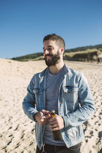 Young man opening bottle while standing on sand against clear sky