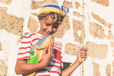 Smiling boy holding fishing rod while standing against wall