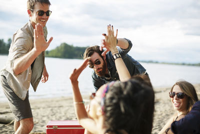 Happy friends doing high-five while enjoying at riverbank against cloudy sky