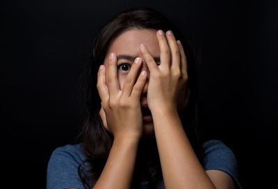 Portrait of scared young woman hiding face standing against black background