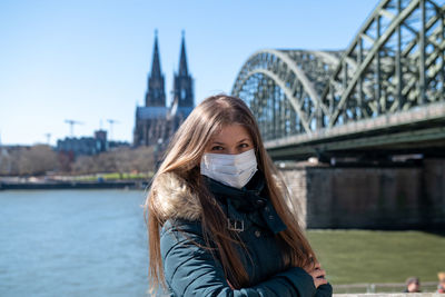 Portrait of young woman wearing mask standing against bridge over river