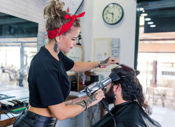 Side view of stylish female master in black leather pants drying hair of bearded male client while working in barbershop