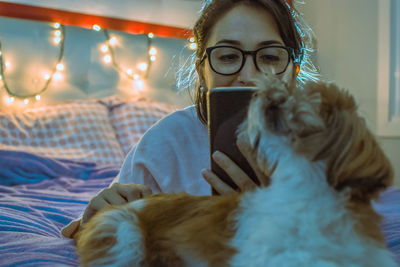 Smiling young woman using phone by dog relaxing on bed at home