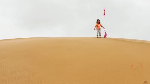 Full length of girl throwing shovel while standing on sand dune against sky