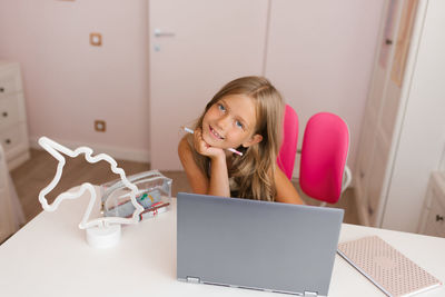 Caucasian cute schoolgirl studies at home using a laptop, sits at her desk, looks at the camera