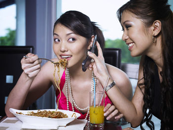 Female friends eating spaghetti while talking on mobile phone in restaurant