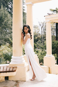 A beautiful brunette lady in an elegant wedding dress poses among the columns in the old city park