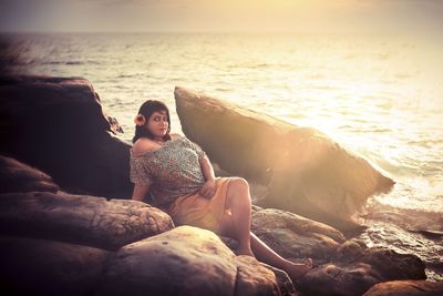 Woman sitting on rock at beach against sky during sunset