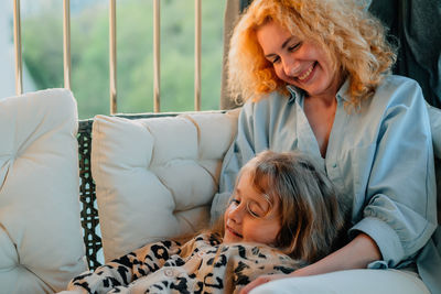 A small, happy girl, a child on her mother's lap with a smile on the sofa at home at sunset 