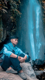 Young man looking away while sitting on rock in forest