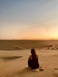 Woman girl sitting on sand in desert against sky during sunset