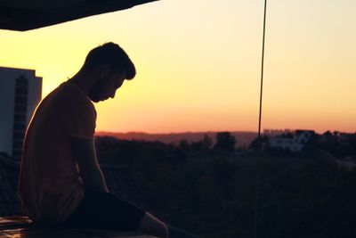 Side view of young man sitting against sky during sunset