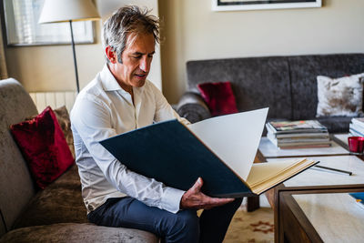 Aged cheerful happy male reviewing business plan while resting on soft comfortable sofa near table with papers