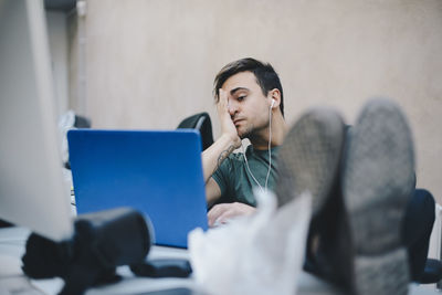 Tired computer programmer using laptop while sitting with feet up at desk in office