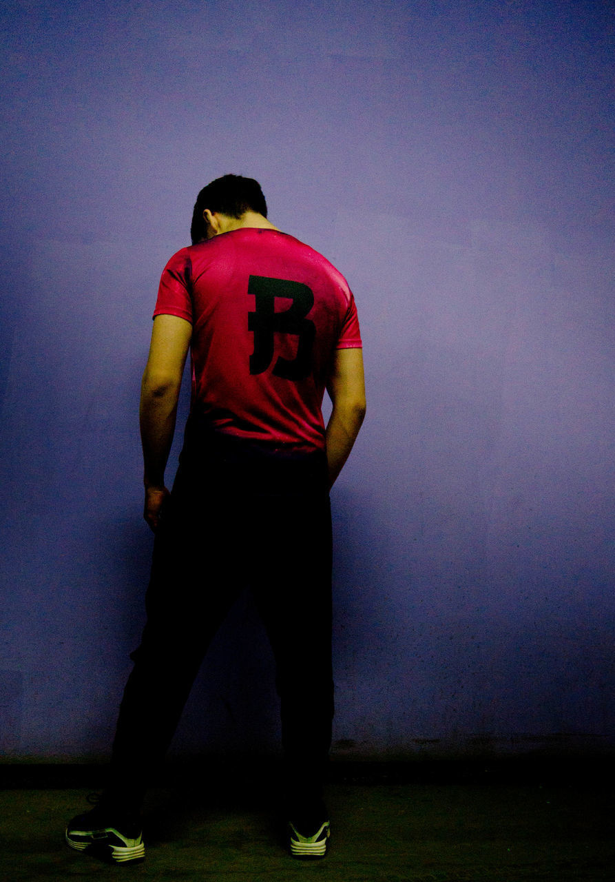 REAR VIEW OF MAN STANDING AGAINST WALL IN ILLUMINATED ROOM